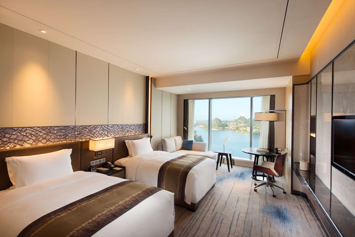 Habitación de hotel con dos camas, un escritorio con silla, televisor y ventana grande con vista a un cuerpo de agua.