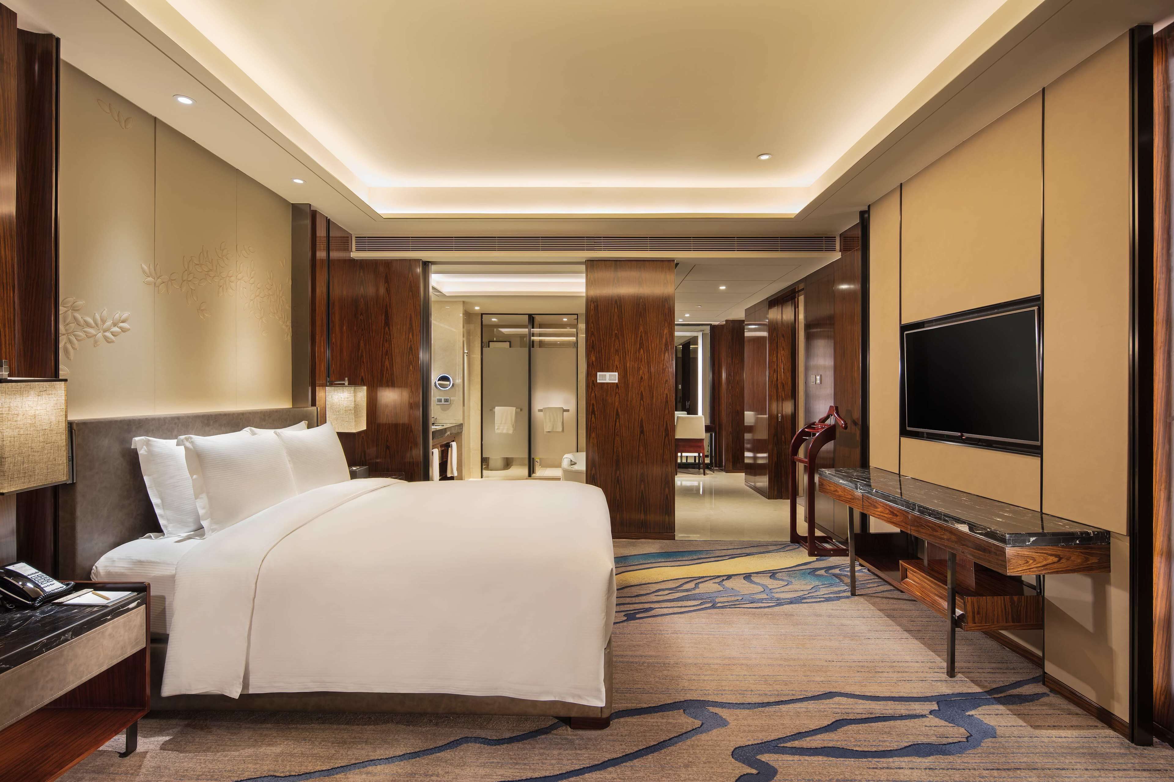 A hotel room with a large bed, a desk with a flat-screen TV, a bathroom, and a hallway leading to another room.