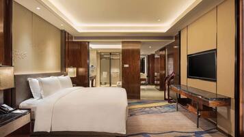 A hotel room with a large bed, a desk with a flat-screen TV, a bathroom, and a hallway leading to another room.