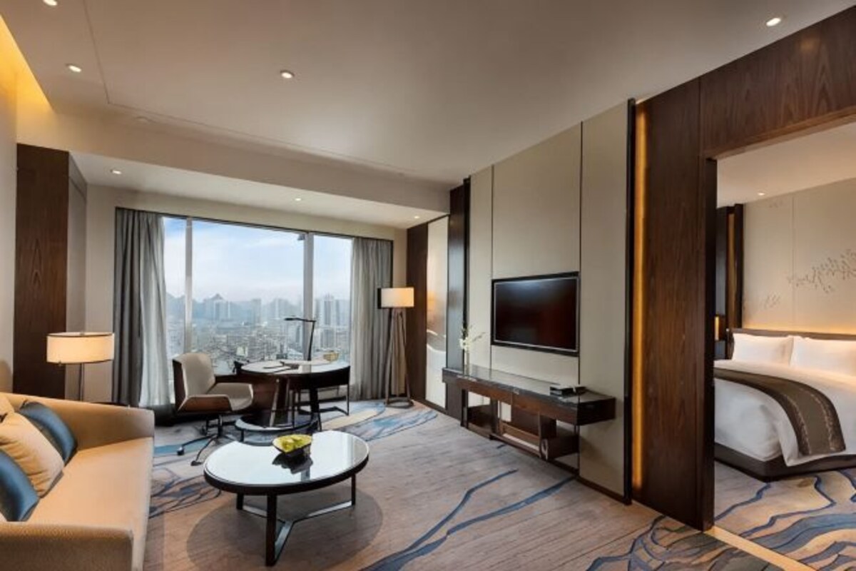 A hotel room with a city view, a flat-screen TV, a bed, a sofa, a coffee table, and a dining area.