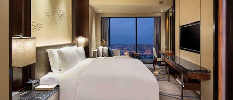 A hotel room with a large bed, a desk with a flat-screen TV, a city view, and a modern design.