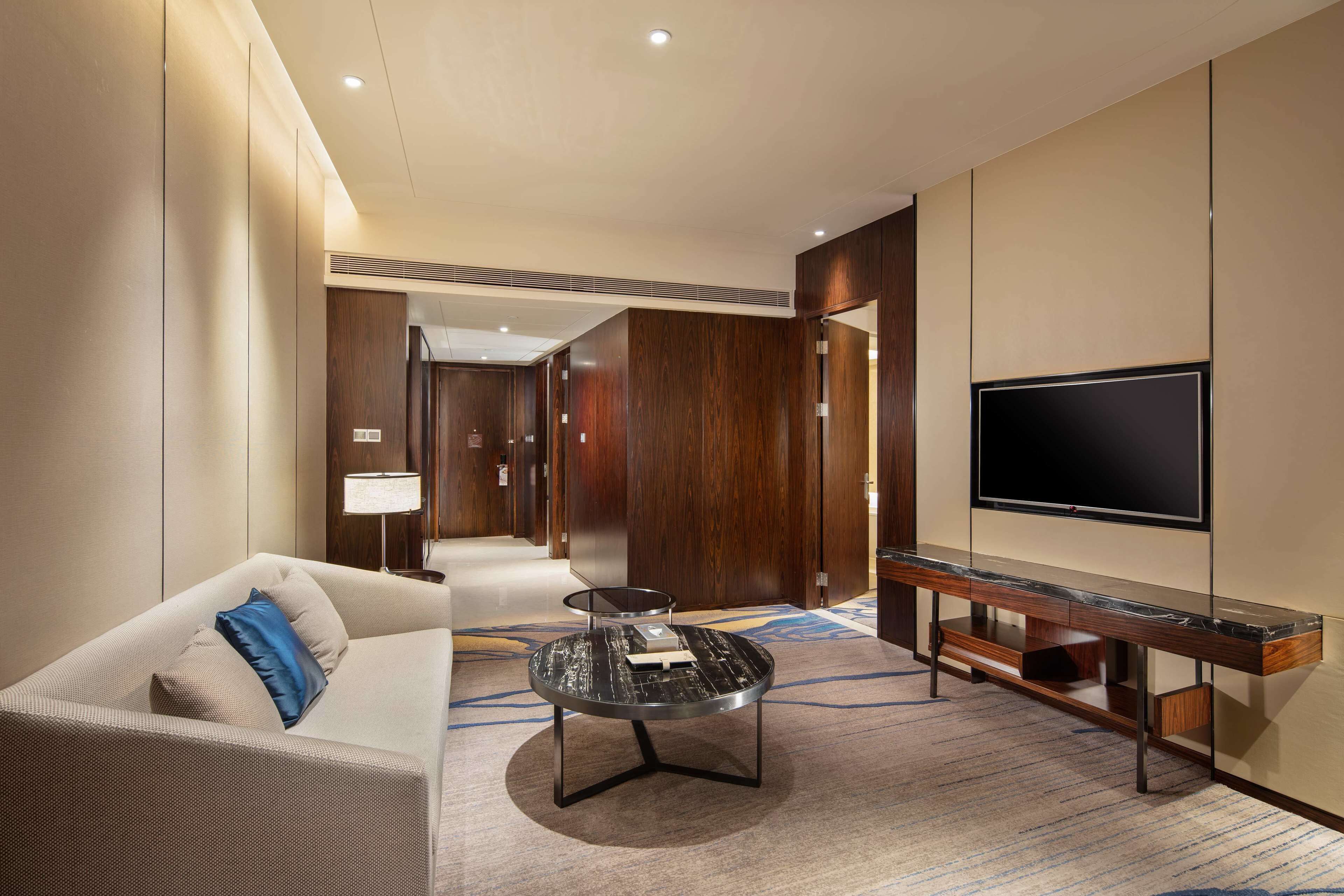 A modern hotel room with a sofa, a coffee table, a TV, and a carpeted floor.