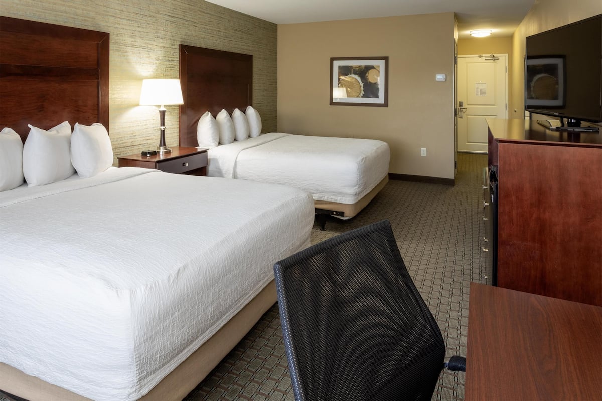 A hotel room with two beds, a desk, a chair, a lamp, a painting, and a television.