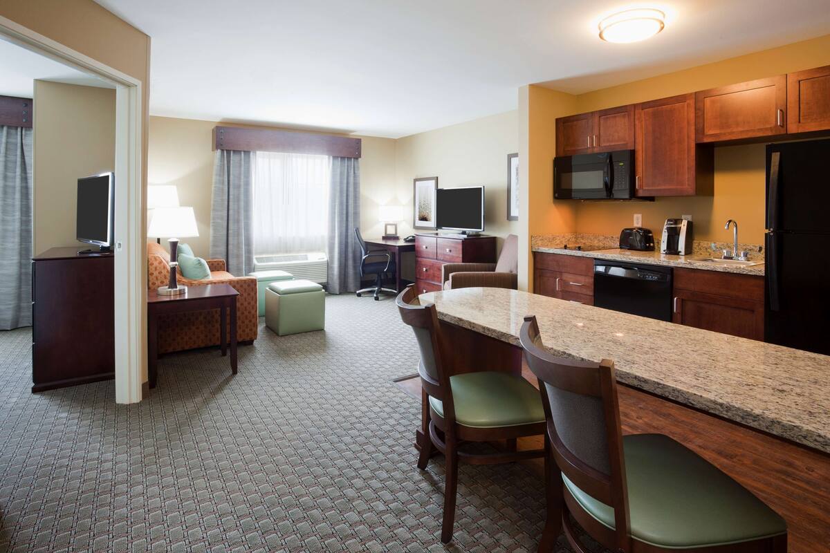 A hotel room with a kitchenette, a dining area with a table and chairs, a TV, and a window with curtains.