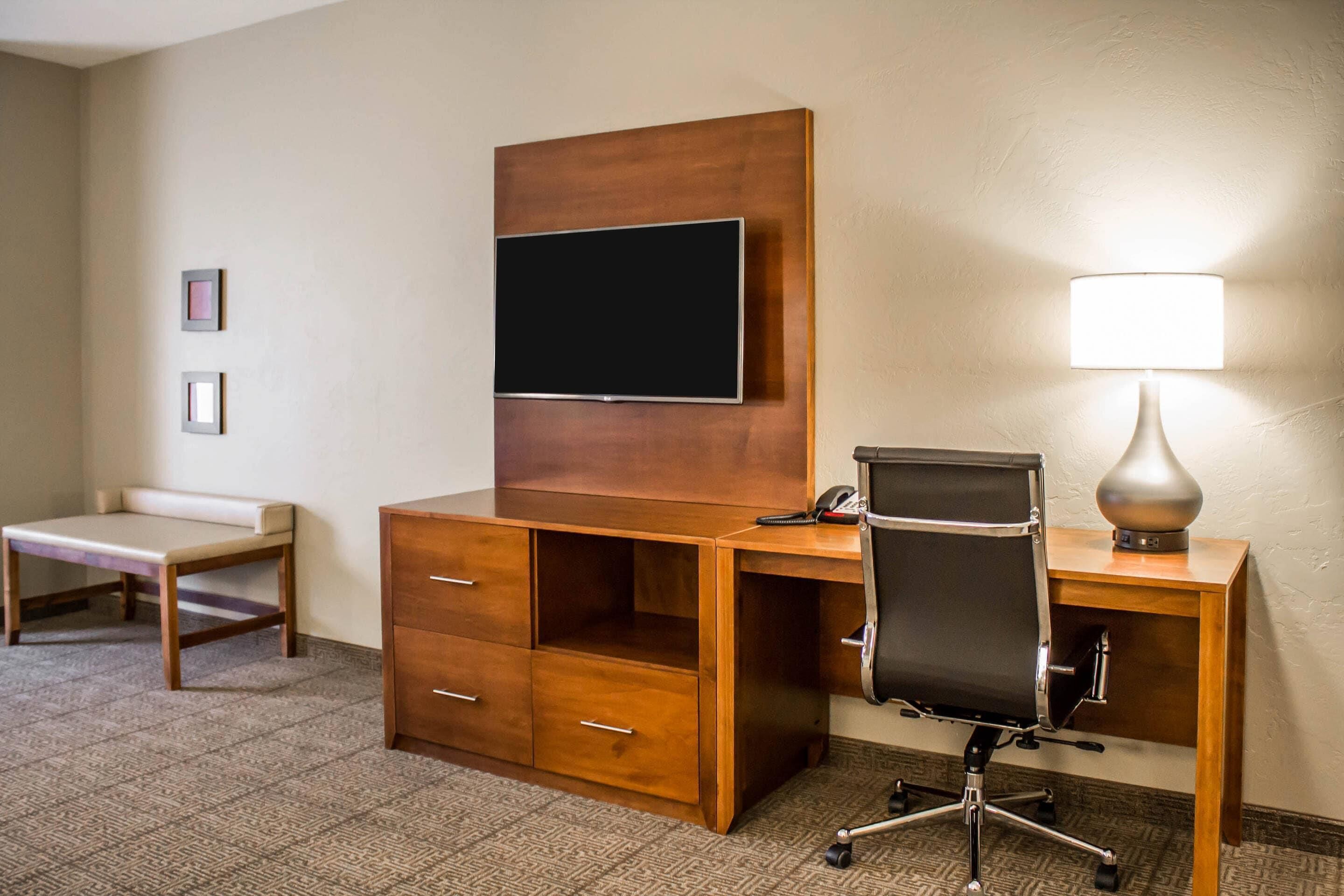 A hotel room with a wooden desk, a chair, a television mounted on the wall, a lamp, and a small table.