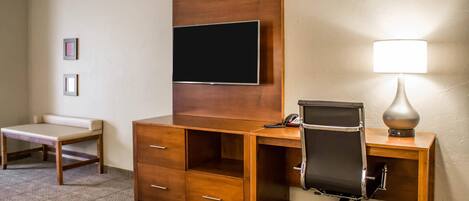 A hotel room with a wooden desk, a chair, a television mounted on the wall, a lamp, and a small table.