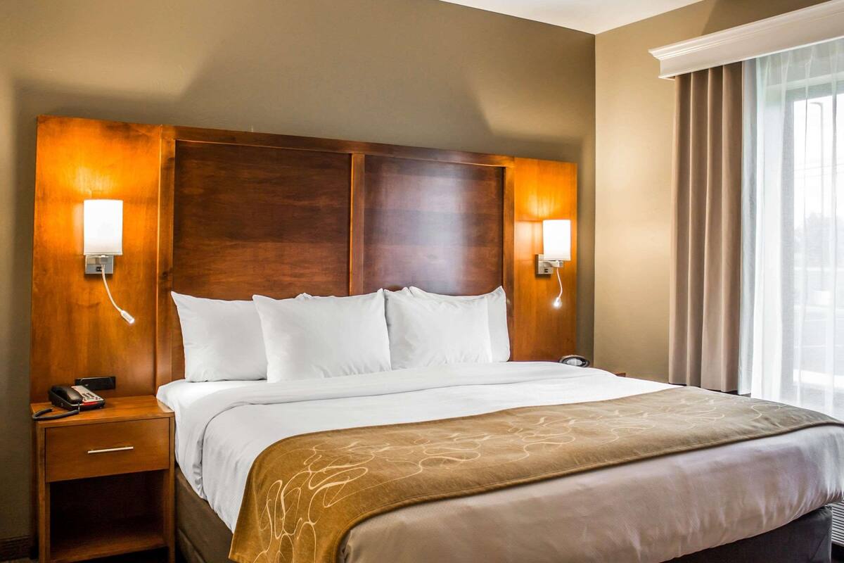 Een hotelkamer met een groot bed, houten hoofdbord, bedlampen en een raam met gordijnen.