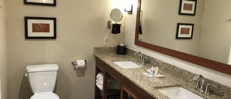 Une salle de bain comprenant des toilettes, deux lavabos, un grand miroir et des œuvres d’art encadrées accrochées aux murs.