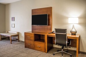 A hotel room with a wooden desk, a chair, a television mounted on the wall, a lamp, and a small bench. - Comfort Suites Hartville - North Canton (Hartville)