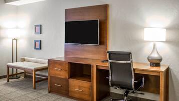 A hotel room with a wooden desk, a flat-screen TV mounted on the wall, a chair, a bedside table, and a lamp.