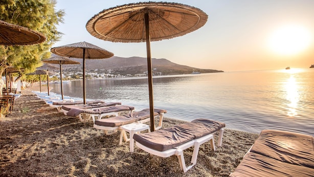A beachside setting with lounge chairs and umbrellas, a calm sea, and a mountain in the background. - Alinda Hotel (Leros)