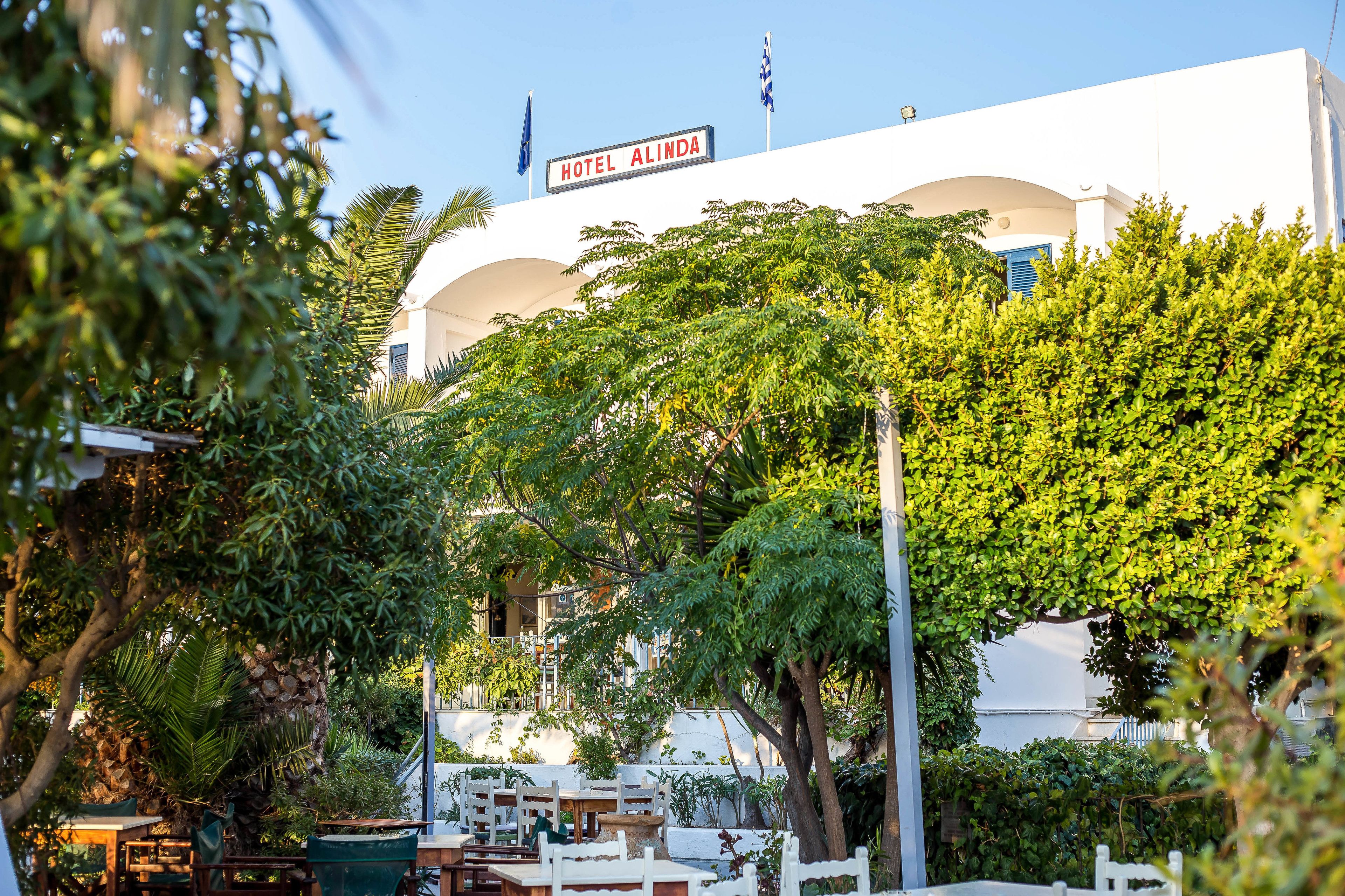A hotel with a white facade, a sign reading "HOTEL ALINDA," and outdoor seating surrounded by lush greenery.