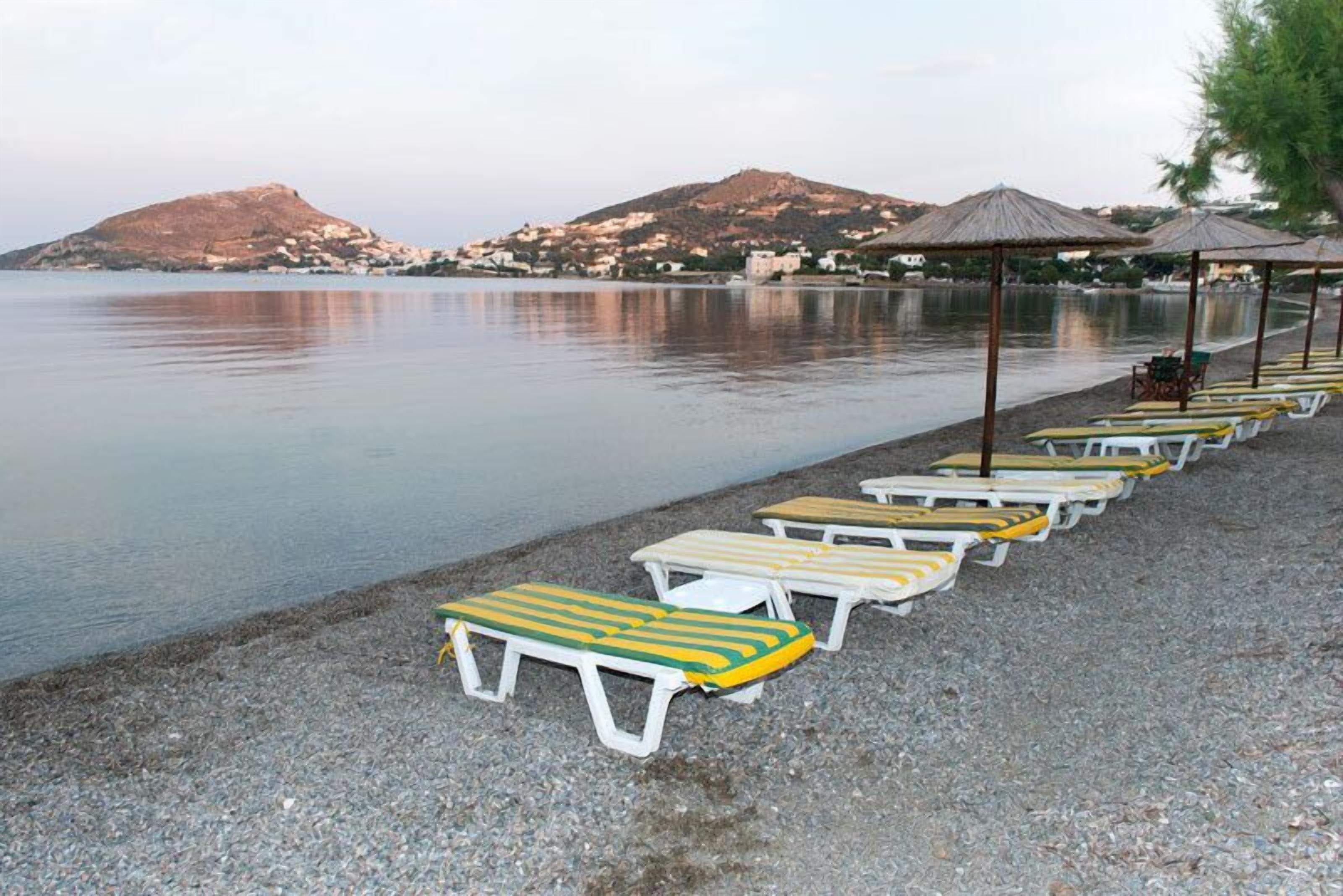 A beach with rows of sun loungers and umbrellas, a calm sea, and hills in the background.