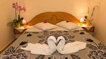 Una cama bien hecha con dos toallas dispuestas en forma de cisne, un cabecero de madera y dos mesitas de noche con lámparas y orquídeas en macetas.