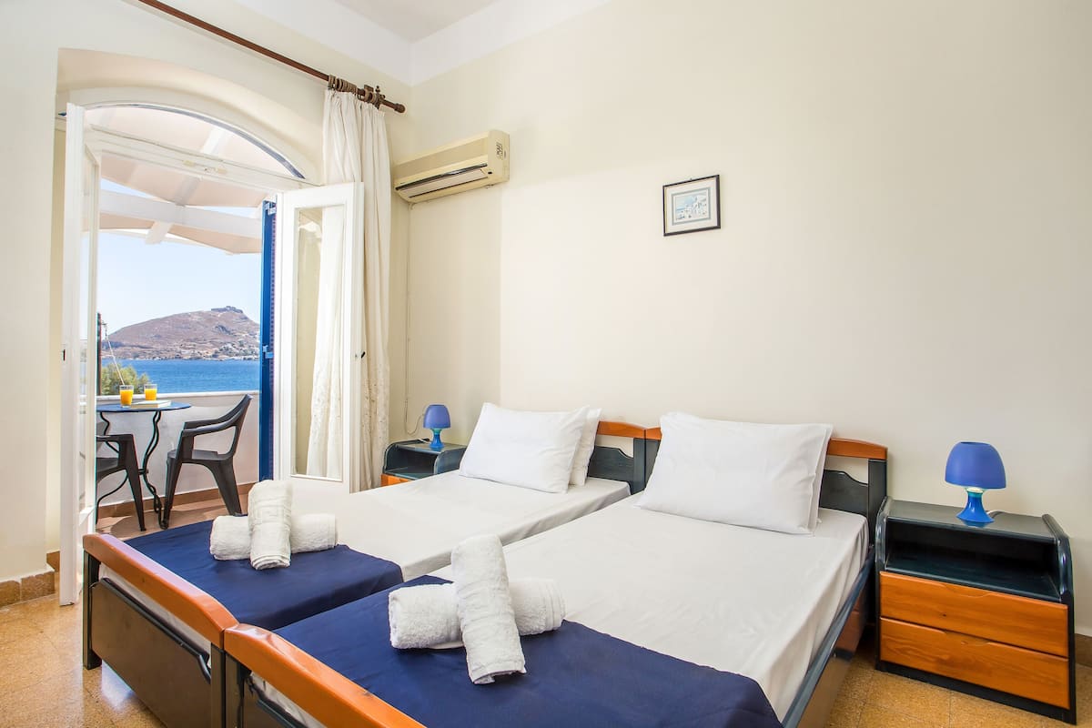 A hotel room with two beds, a small table with chairs, a nightstand with a lamp, and a view of the sea and a mountain.