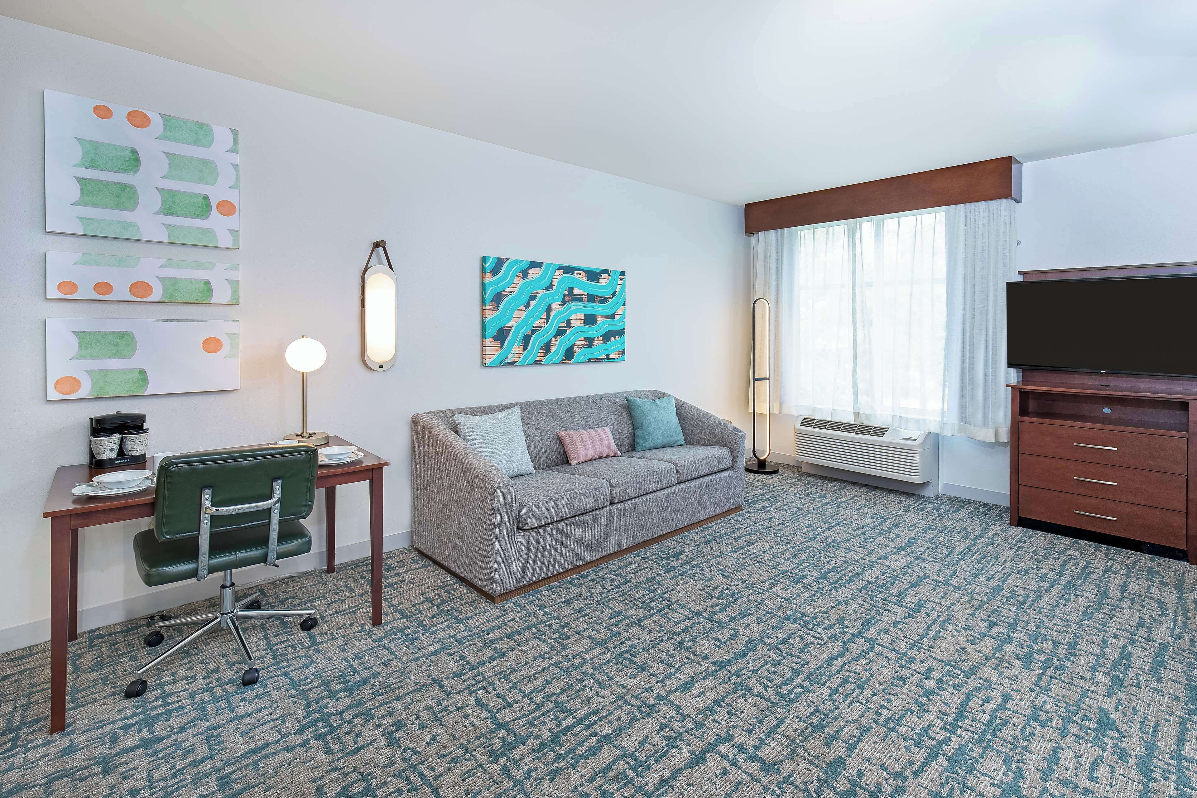 A hotel room with a grey sofa, a desk with a chair, a flat-screen TV, and artwork on the walls.