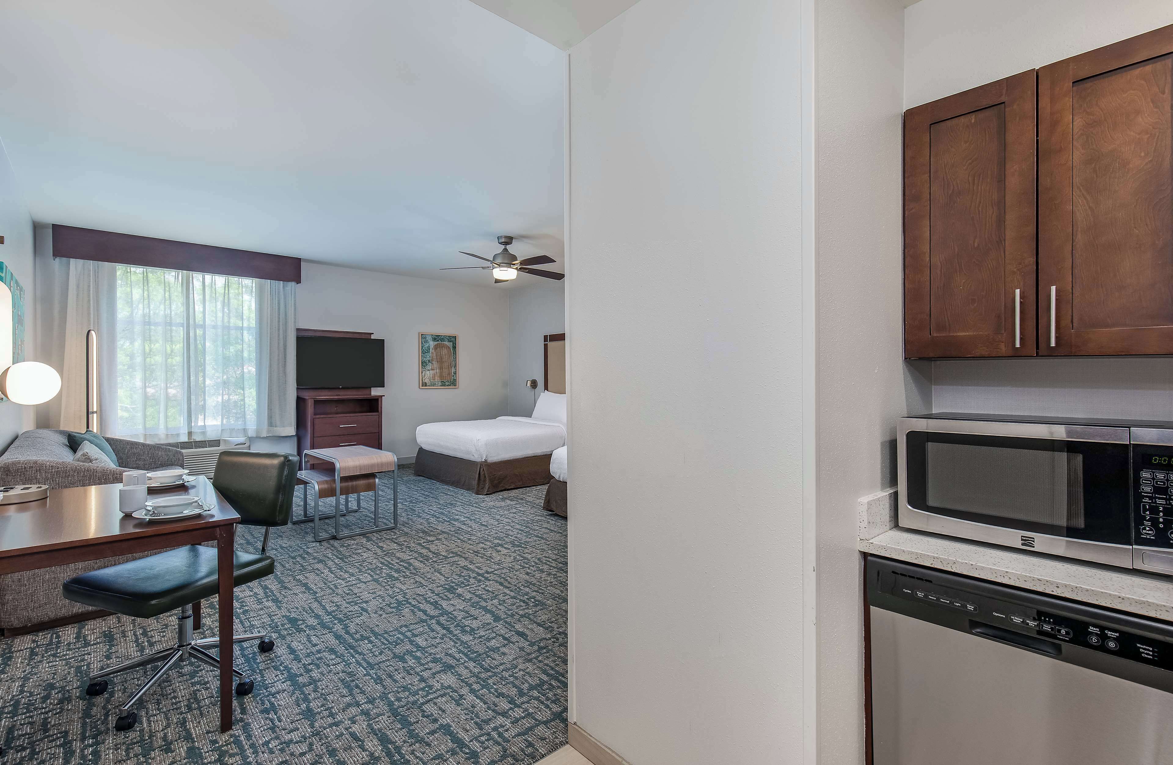 A hotel room with a kitchenette, a bed, a desk with a chair, a television, and a ceiling fan.