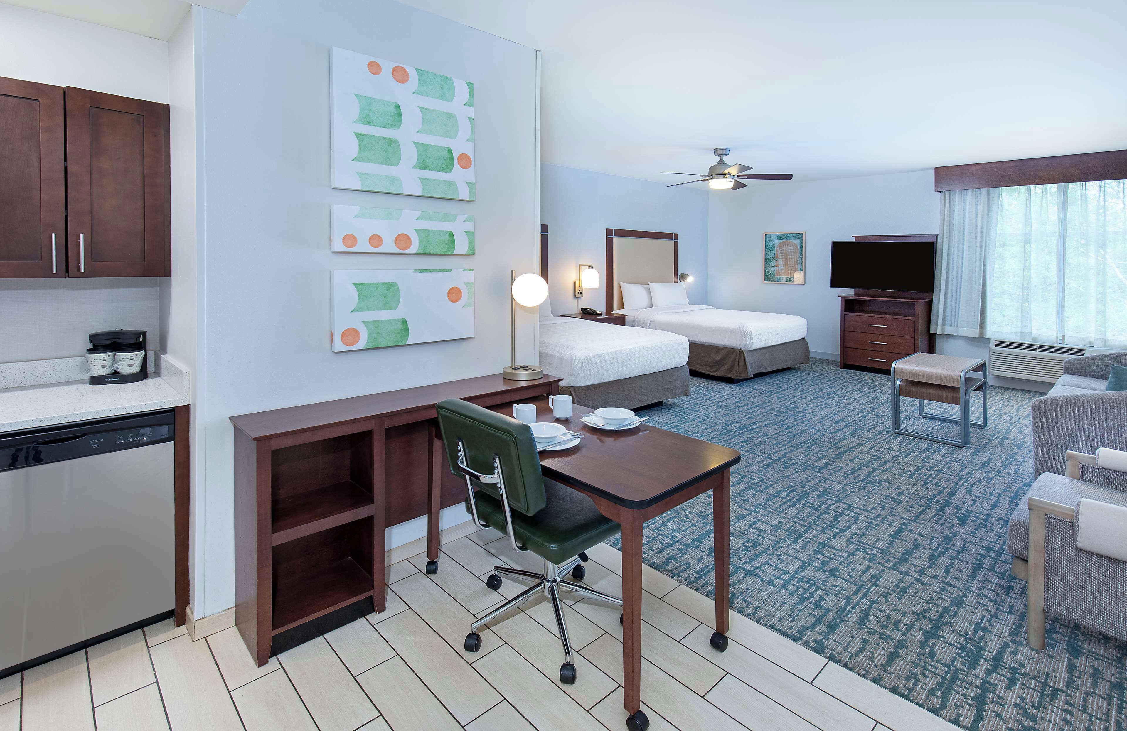 A hotel room with a kitchenette, two beds, a desk with a chair, a TV, and a large window.