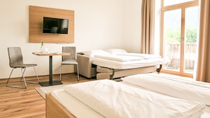 Habitación de hotel con cama, escritorio con dos sillas, televisor montado en la pared y un ventanal con cortinas.