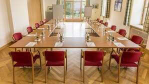 Un salón de conferencias con una mesa larga, sillas rojas, caballetes con pizarras blancas y vistas al exterior con vegetación.