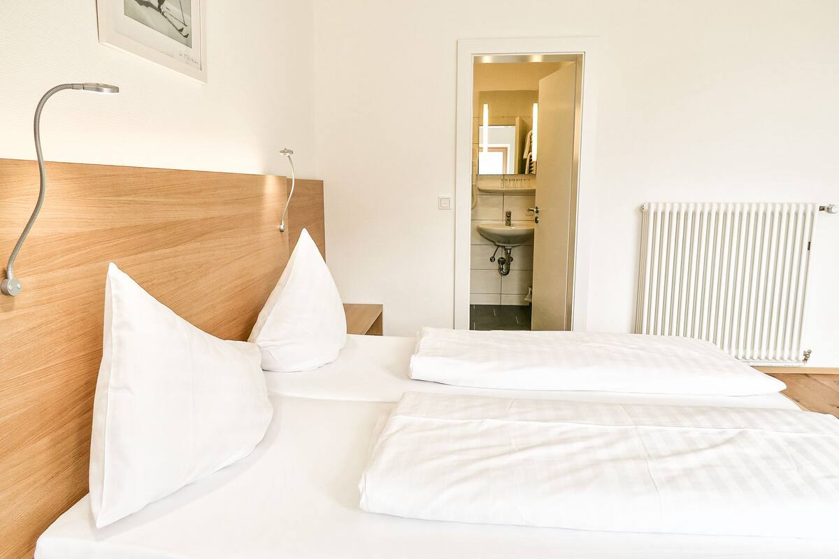 Une chambre d’hôtel avec une tête de lit en bois, un lit avec du linge de maison blanc, une douche avec un pommeau de douche amovible et un radiateur.