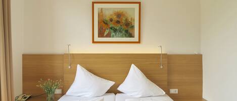 Una cama doble con sábanas blancas, cabecera de madera, mesita de noche con un florero y un cuadro con girasoles en la pared.
