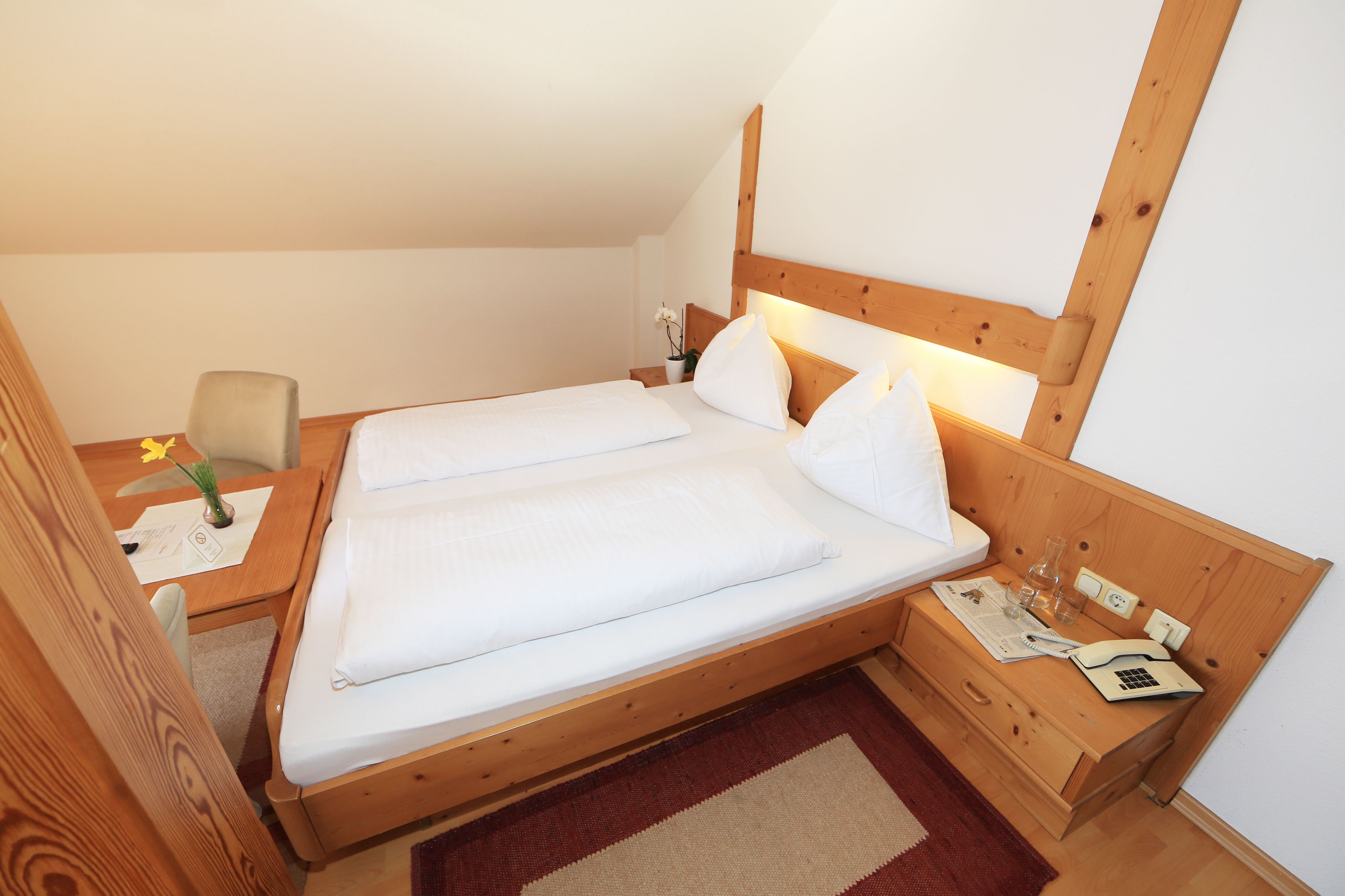 Una cama de madera con ropa de cama blanca, una mesita de noche con una lámpara y un escritorio pequeño con un florero de flores.