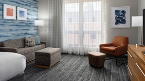 Une chambre d’hôtel avec un tapis gris, un canapé gris, un fauteuil orange, une commode en bois et une grande fenêtre avec des rideaux légers.