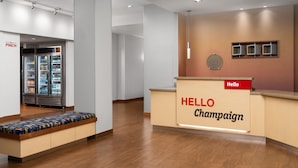Un espace d’accueil d’hôtel comprenant un banc, un réfrigérateur rempli de boissons et une pancarte indiquant « Bonjour, bienvenue à Champaign ».