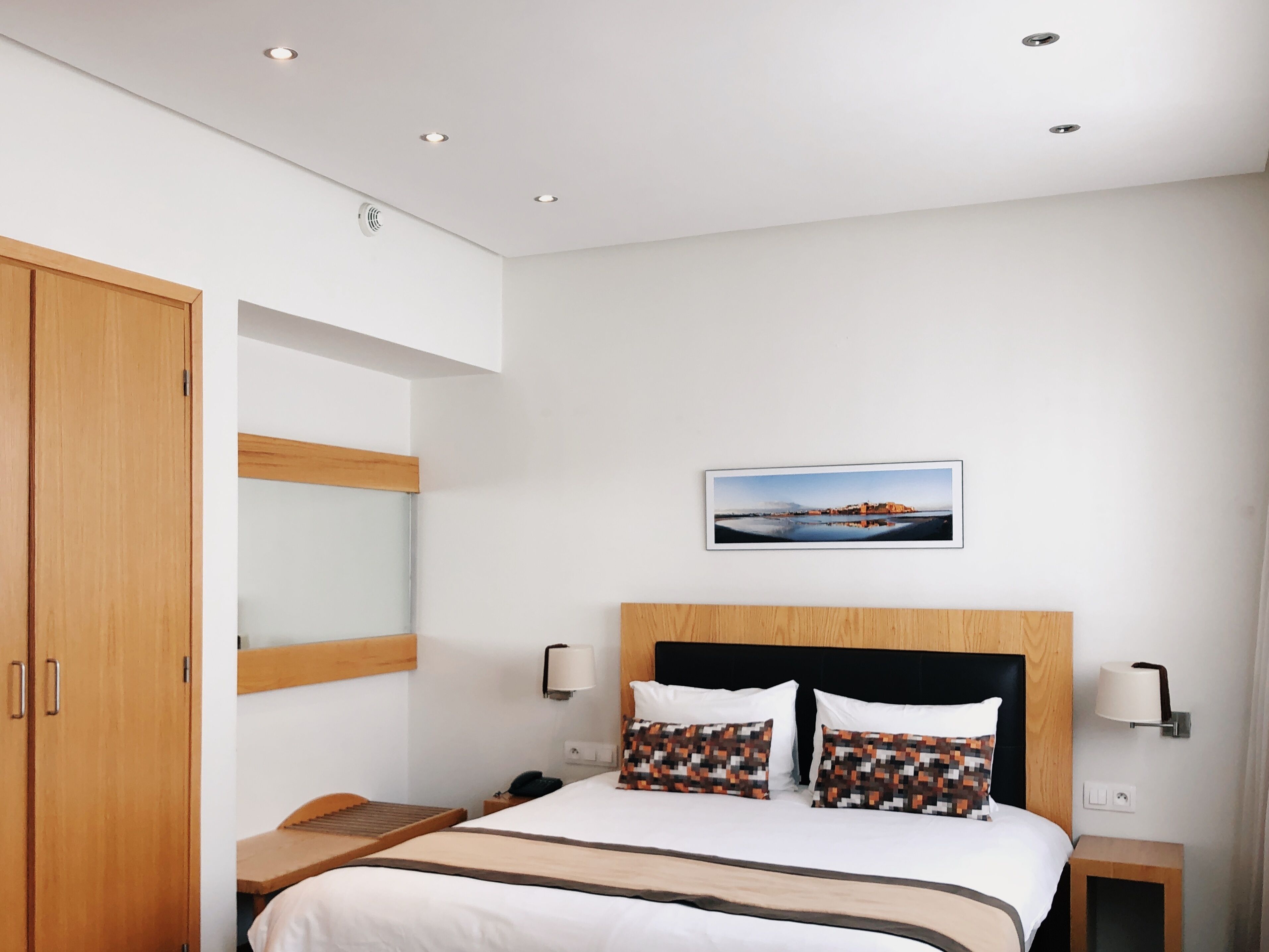 Una habitación de hotel moderna con un armario de madera, una cama con almohadones estampados, una mesita de noche y una lámpara de pared.