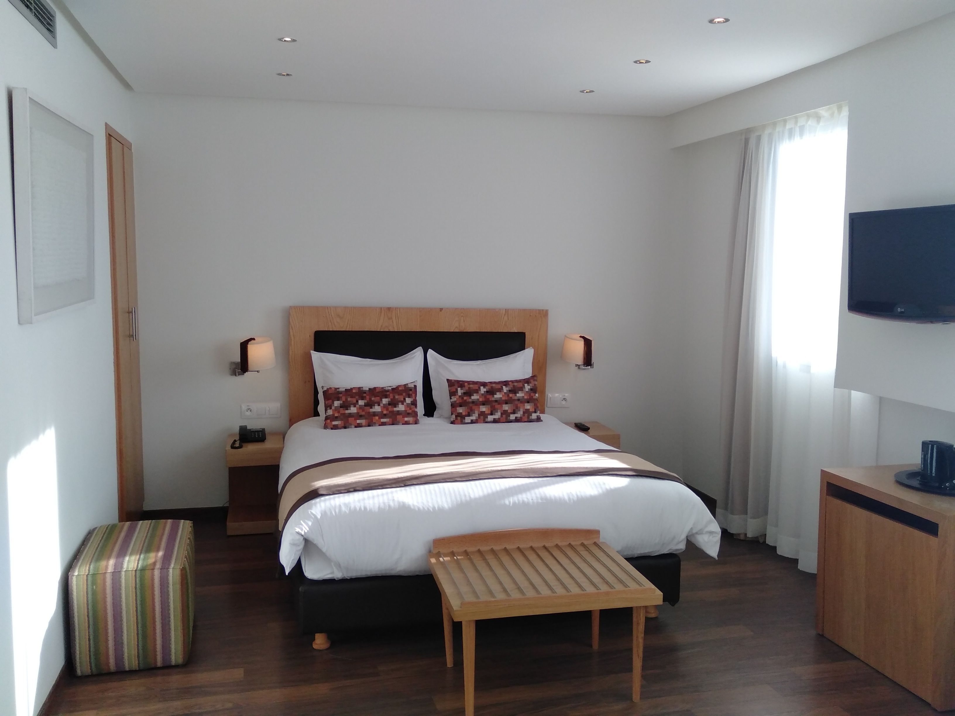 A modern hotel room with a large bed, bedside tables, a TV, and a small wooden bench.