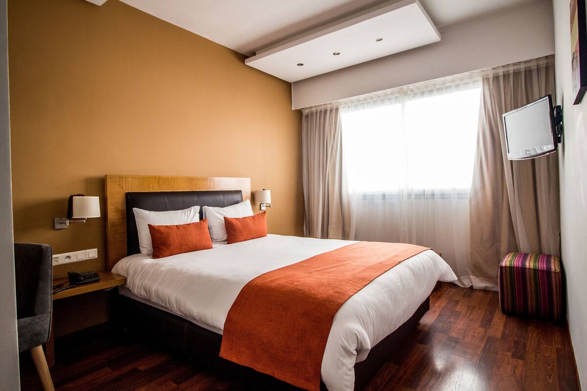A hotel room with a large bed, a desk with a lamp, a television mounted on the wall, and a window with curtains.