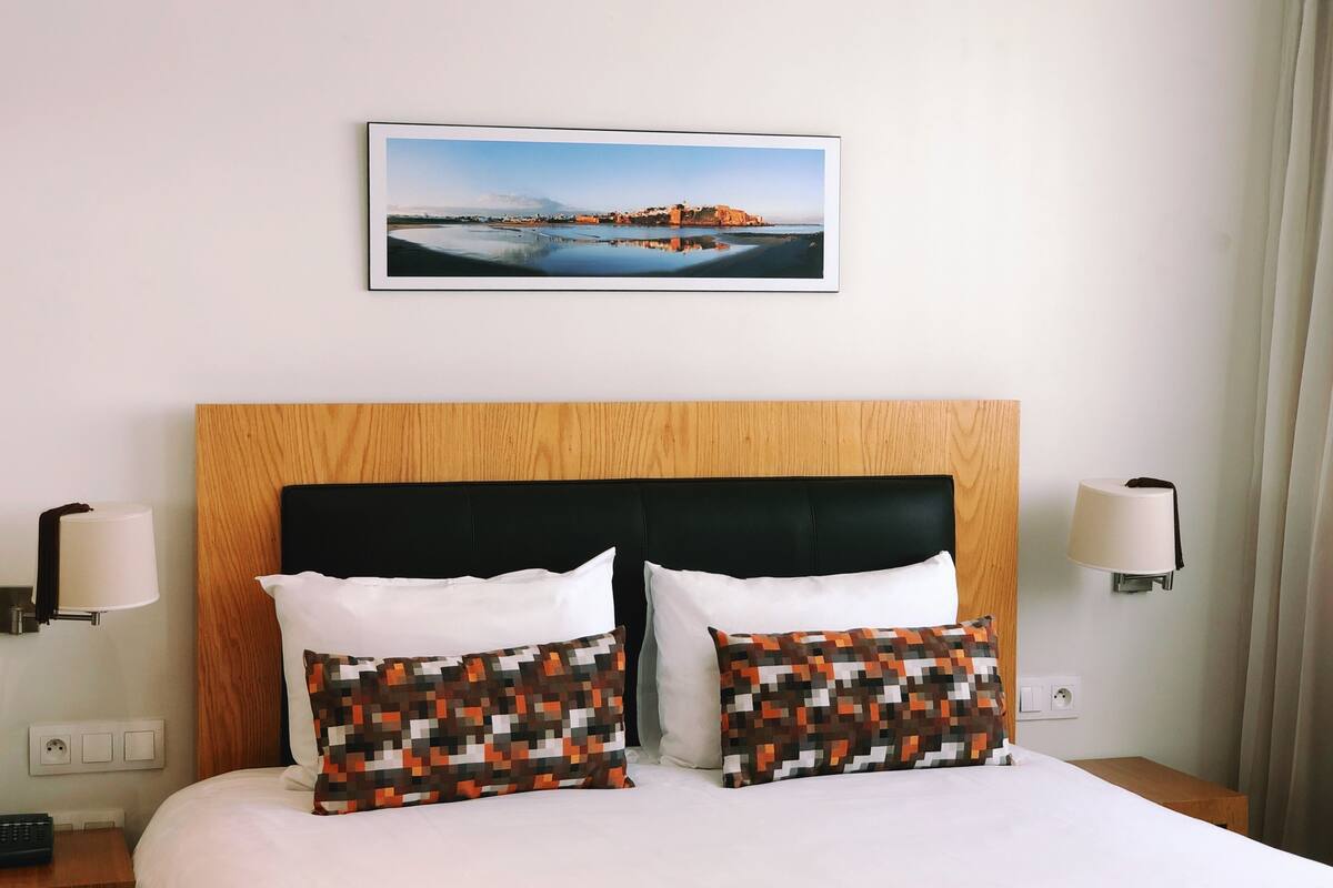 A hotel room with a bed, two pillows, a wooden headboard, a wall-mounted picture, and bedside lamps.