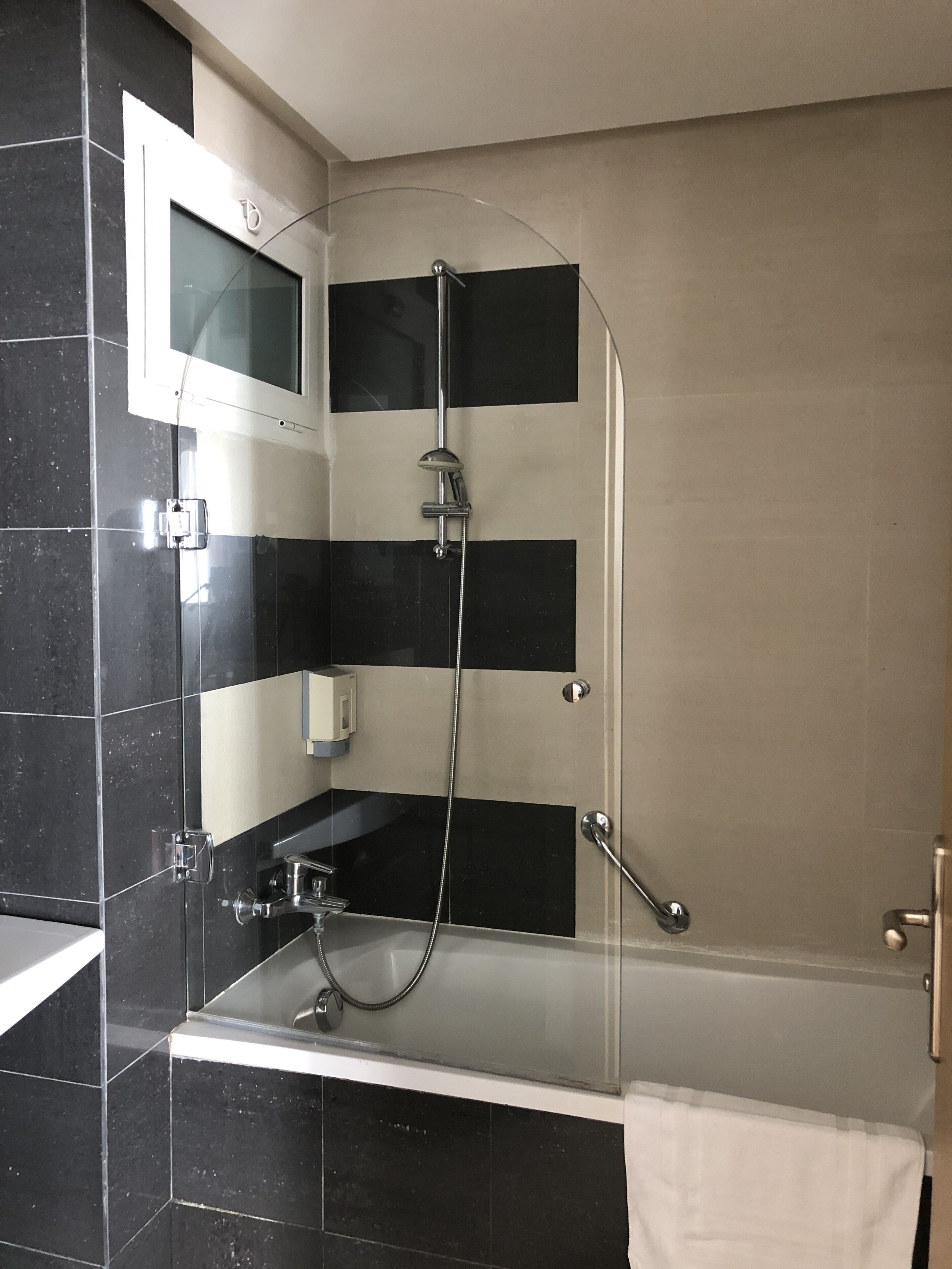 A modern bathroom with a glass-enclosed shower, a white bathtub, and a black and beige tiled wall.