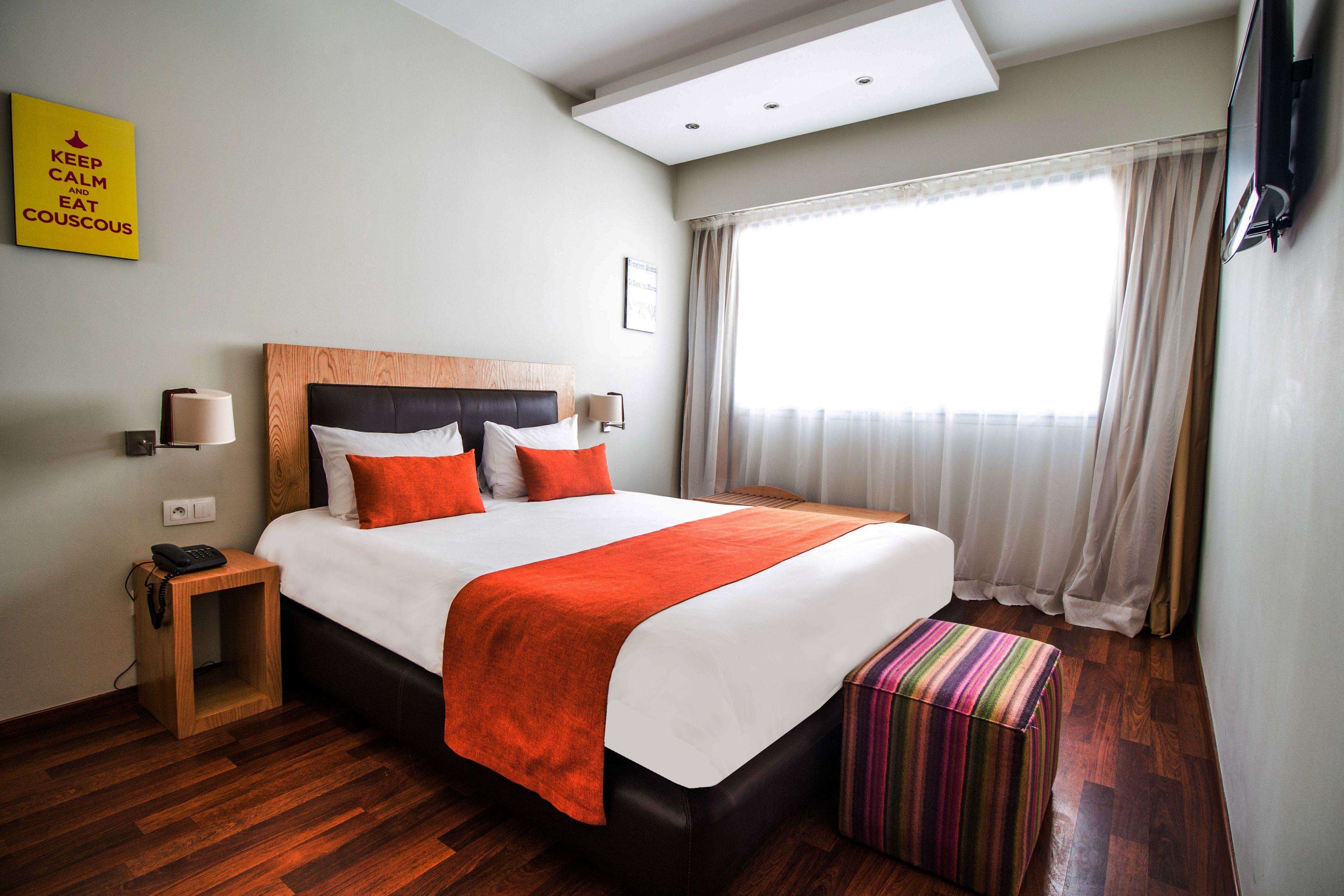 A hotel room with a large bed, a bedside table with a lamp, a small wooden nightstand, a colorful ottoman, and a window with curtains.