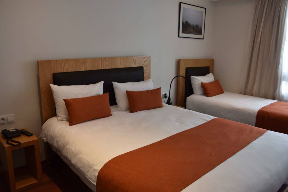 A hotel room with two beds, each with a wooden headboard, white bedding, and orange throw pillows. There is a nightstand with a phone and a lamp.