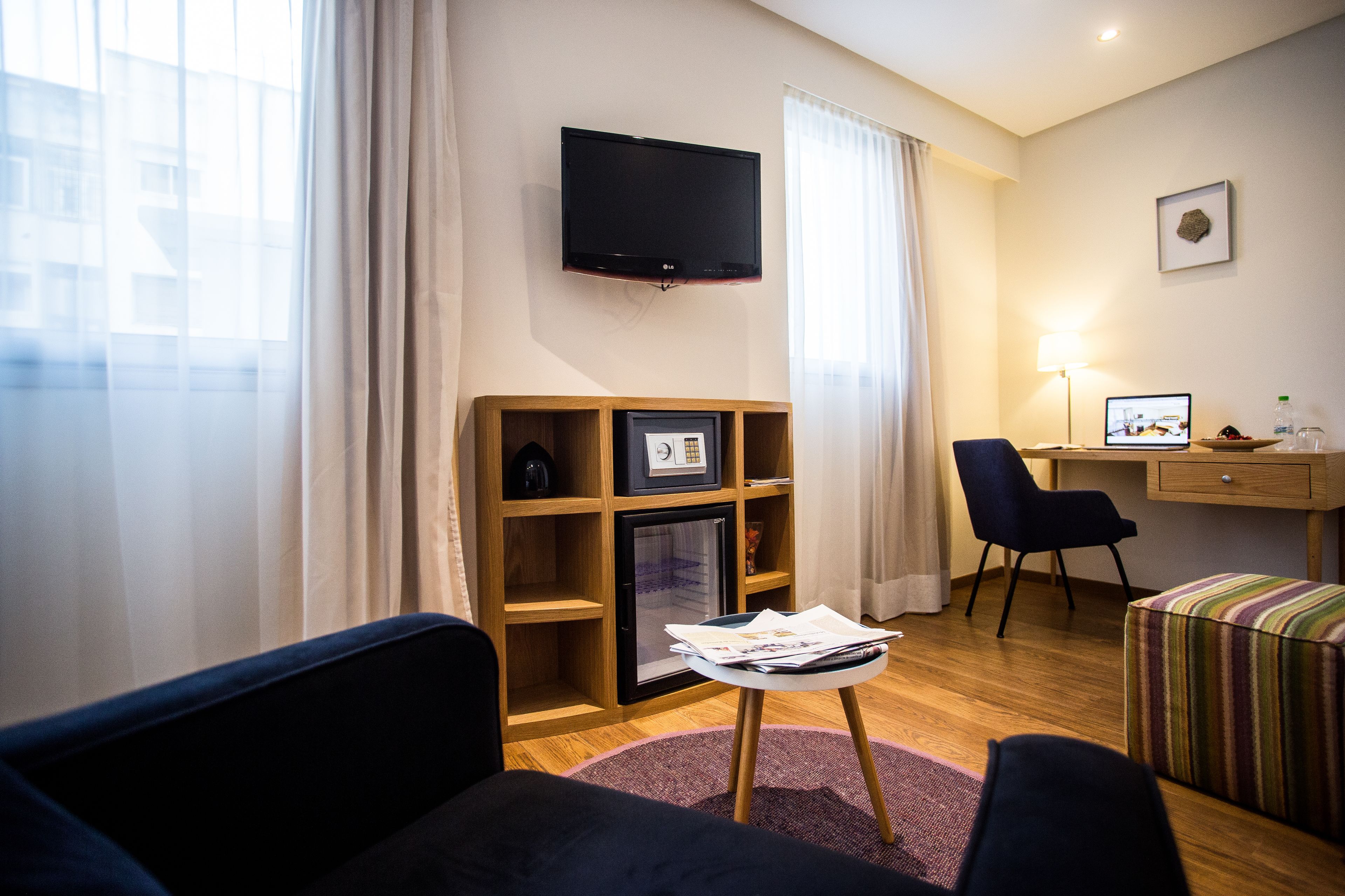 A modern living room with a flat-screen TV, a wooden shelving unit, a small table with magazines, a sofa, a desk with a computer, and a striped ottoman.