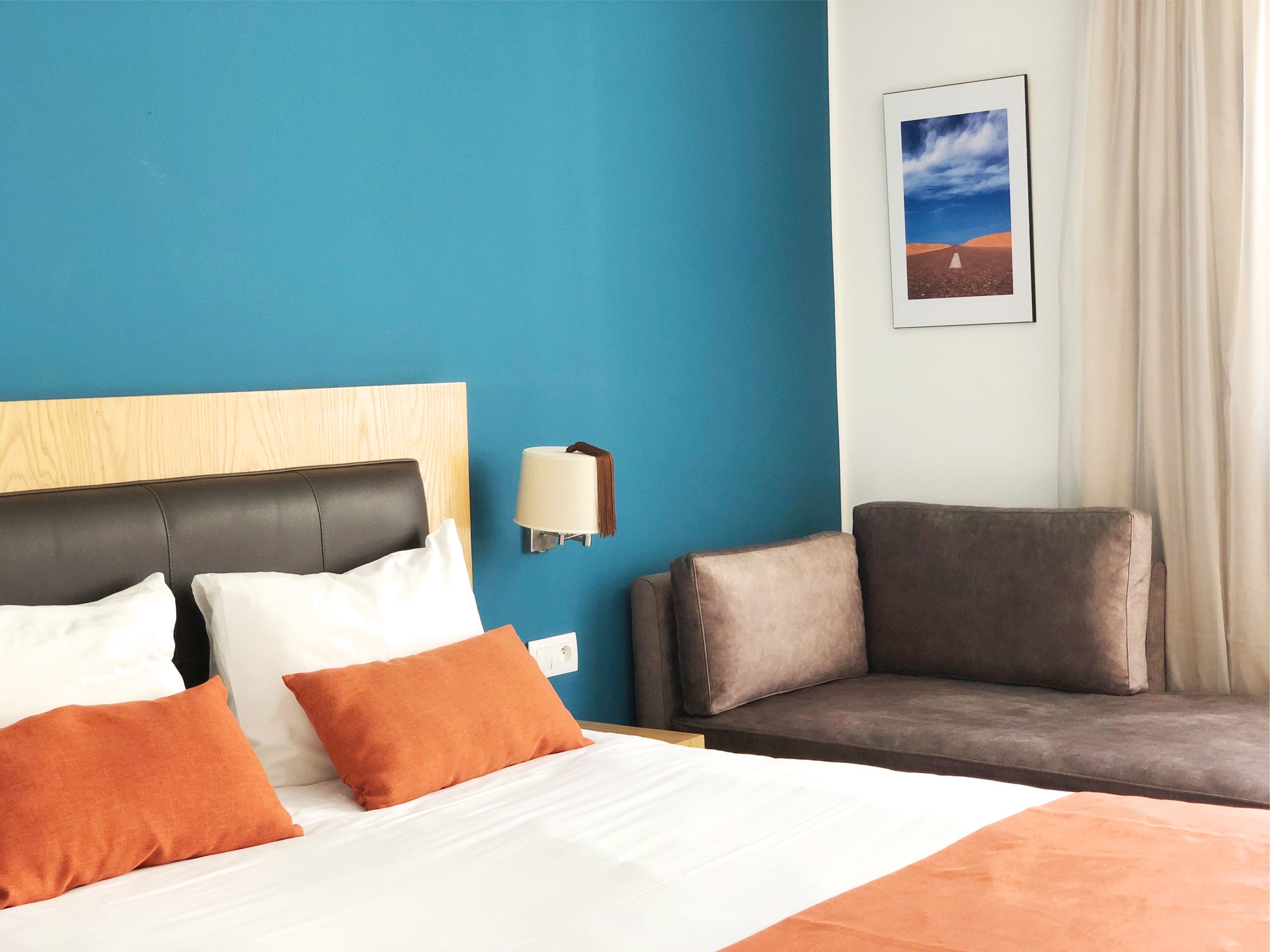 A hotel room with a bed, a headboard, a nightstand, a lamp, a couch, and a framed picture on the wall.