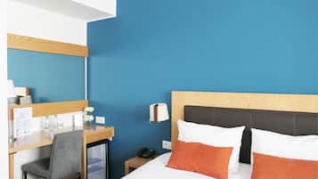 Hotelkamer met een bed, een bureau, een stoel, een kleine koelkast en een nachtkastje met een lamp en telefoon.