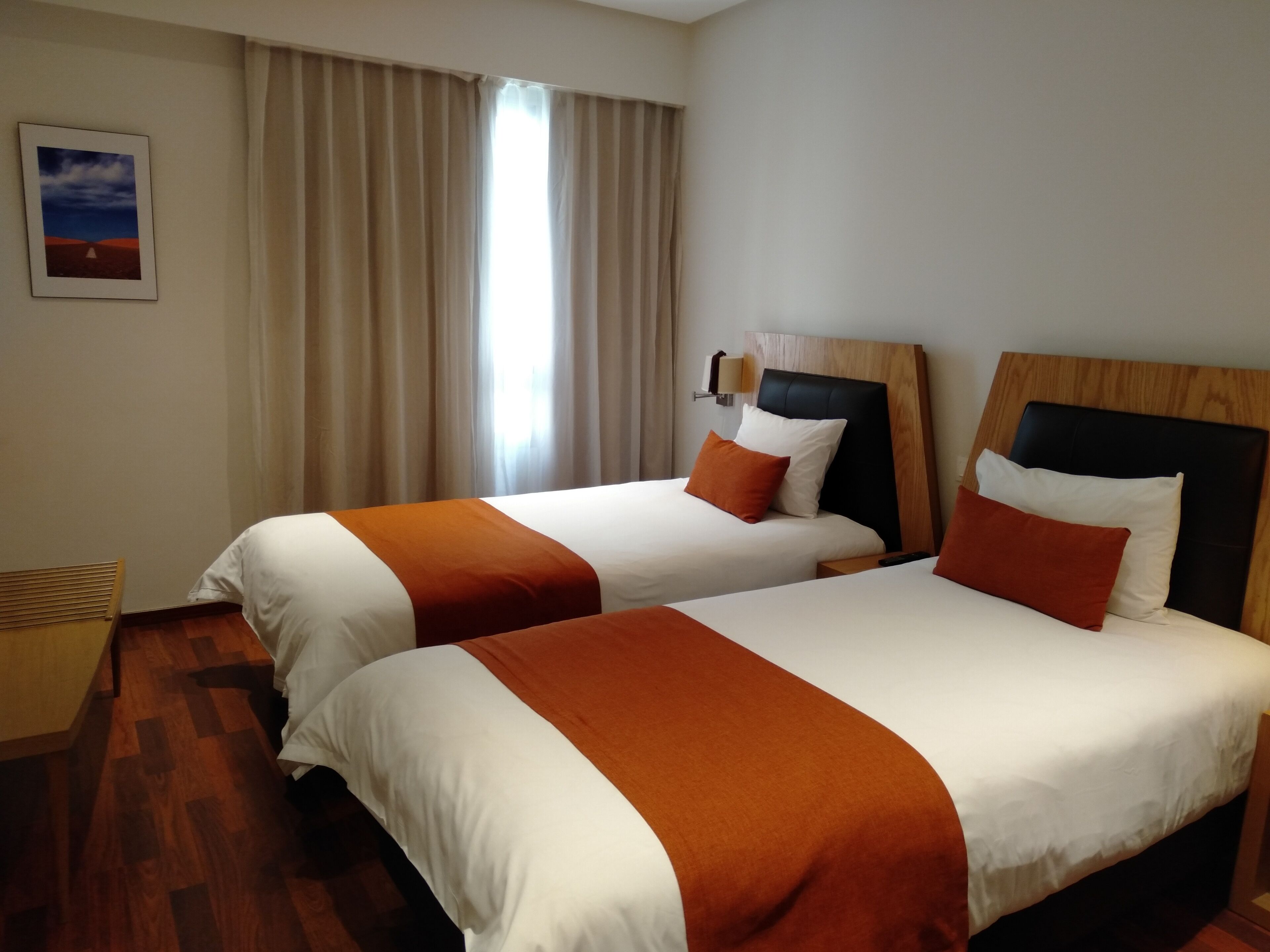 Quarto de hotel com duas camas, cortinas bege, um quadro emoldurado na parede e chão de madeira.