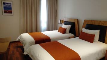 Quarto de hotel com duas camas, cortinas bege, um quadro emoldurado na parede e chão de madeira.