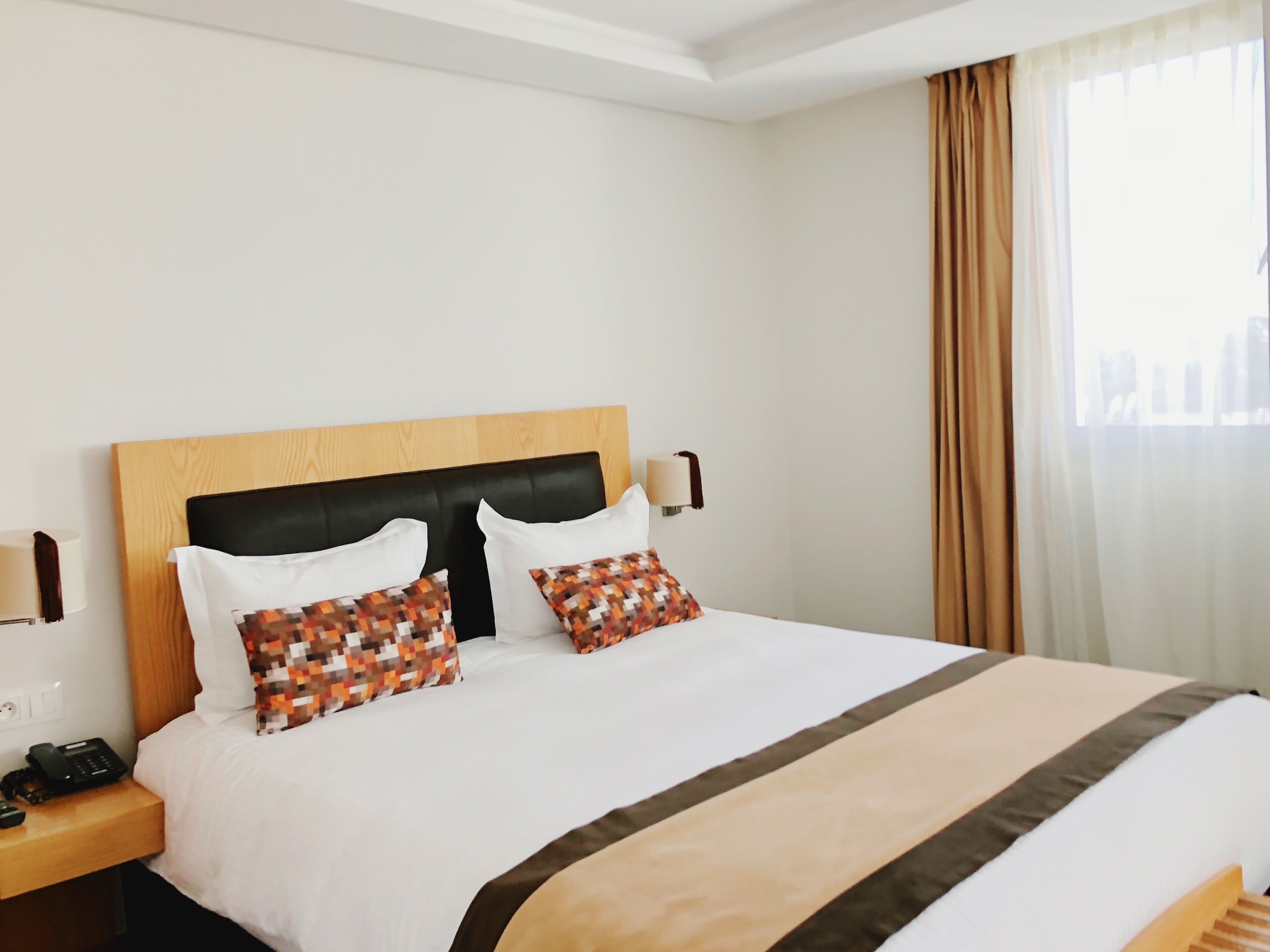 A hotel room with a large bed, a wooden headboard, two white pillows, a patterned pillow, a bedside lamp, and a telephone on the nightstand.