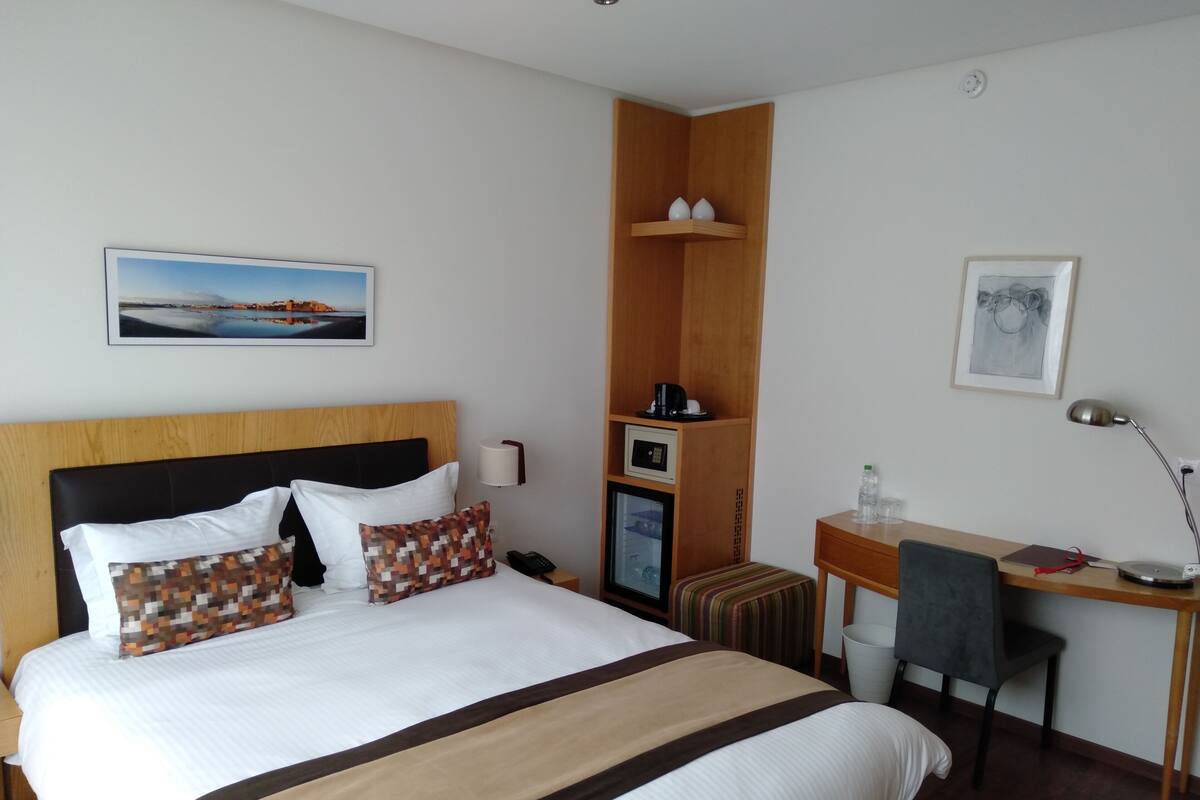 A hotel room with a bed, a desk, a chair, a small cabinet, and a wall-mounted shelf.