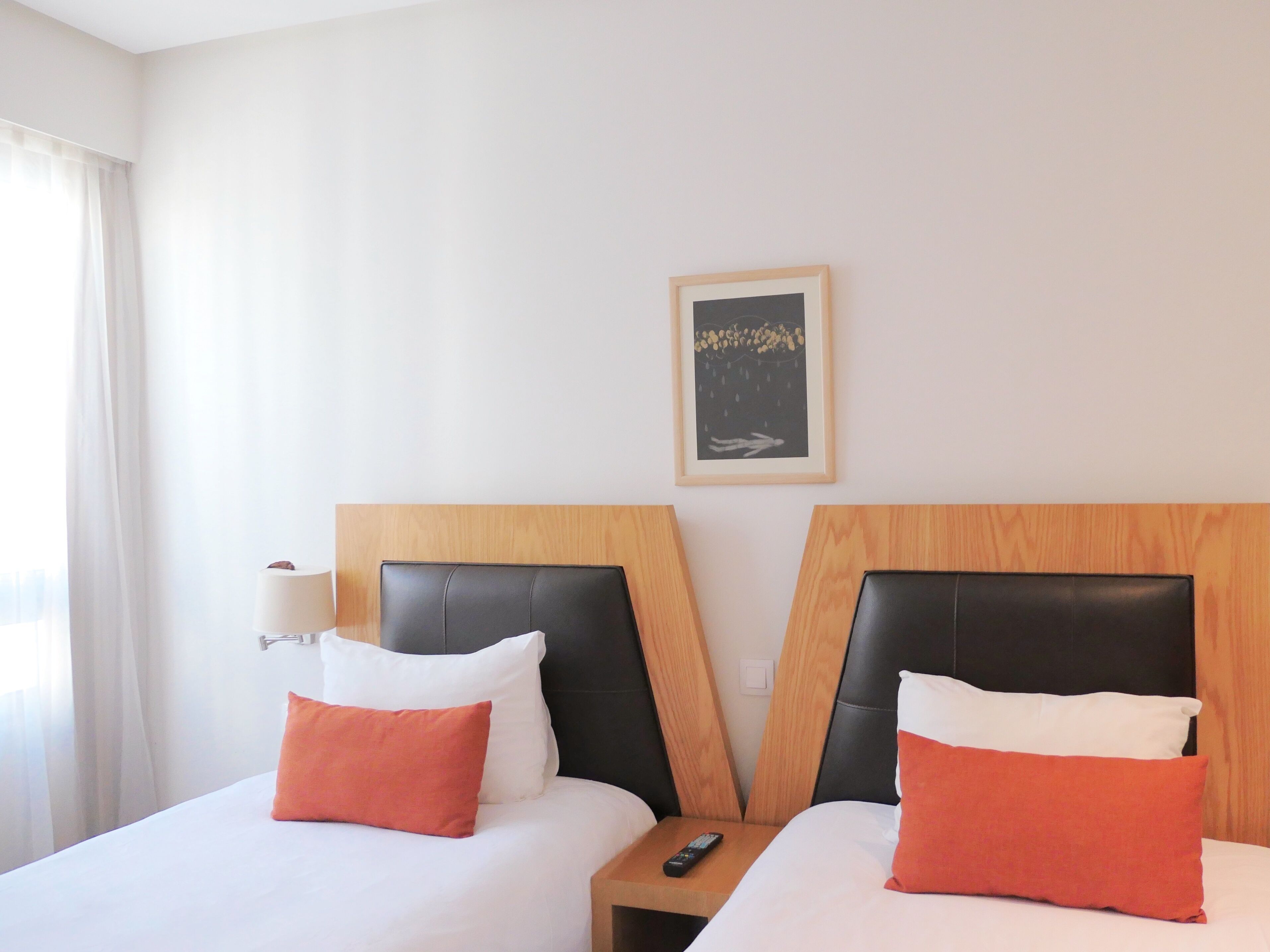 A hotel room with two beds, each with a wooden headboard, white bedding, and orange pillows. A remote control is on the nightstand between the beds.
