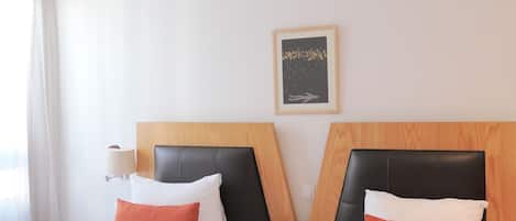 Ett hotellrum med två sängar, var och en med ett träfärgat sänggavel, vita sängkläder och orange kuddar. En fjärrkontroll ligger på nattduksbordet mellan sängarna.
