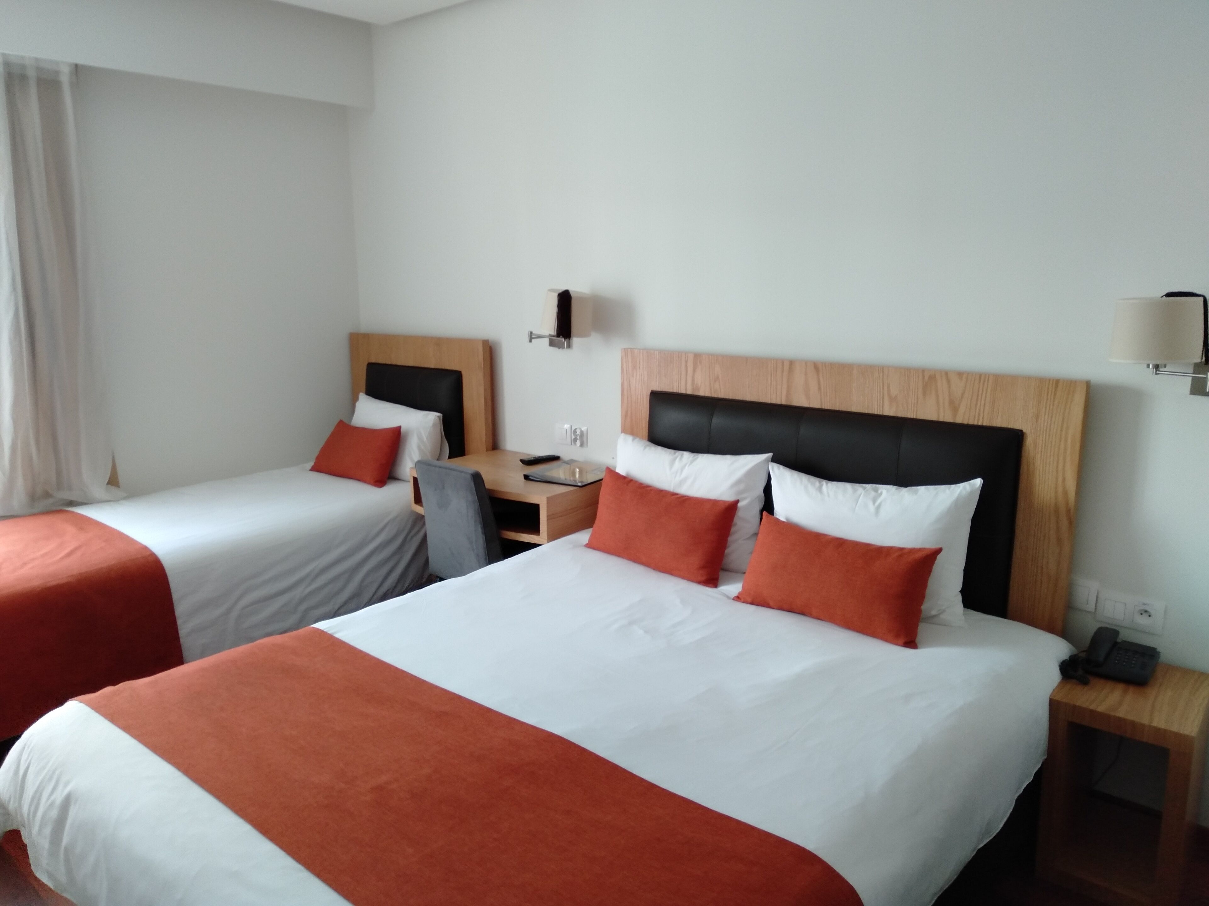 A hotel room with two beds, each with a wooden headboard, white bedding, and red accents. There is a desk with a chair, a lamp, and a phone on the side table.