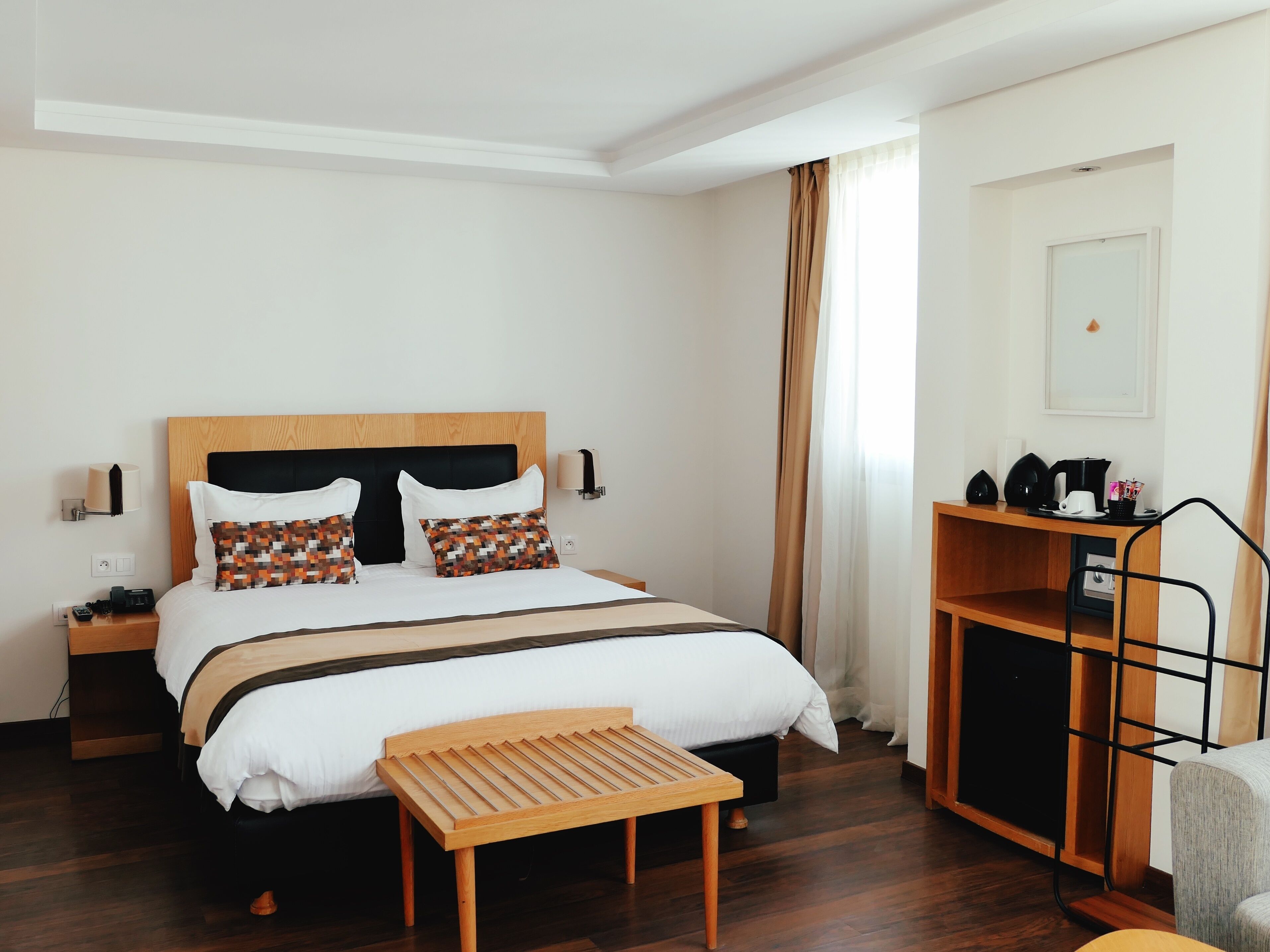 A hotel room with a wooden bed, a bedside table with a lamp, a small wooden bench, and a desk with a coffee maker.