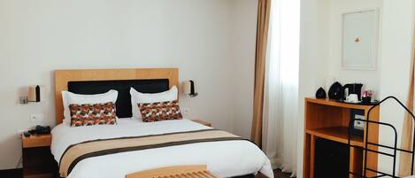 Habitación de hotel con una cama de madera, una mesita de noche con lámpara, un pequeño banco de madera y un escritorio con cafetera.