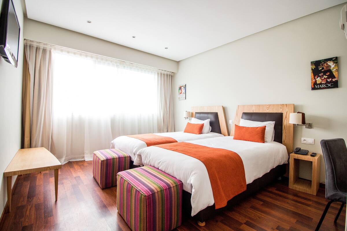 A hotel room with two beds, a wooden table, a striped ottoman, a chair, and a nightstand with a lamp.