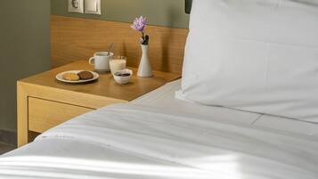 Una cama bien hecha con sábanas blancas, una mesita de noche de madera con un jarrón blanco que contiene una flor morada, un plato con galletas, una taza con cuchara, y un pequeño cuenco con lo que parece mermelada.
